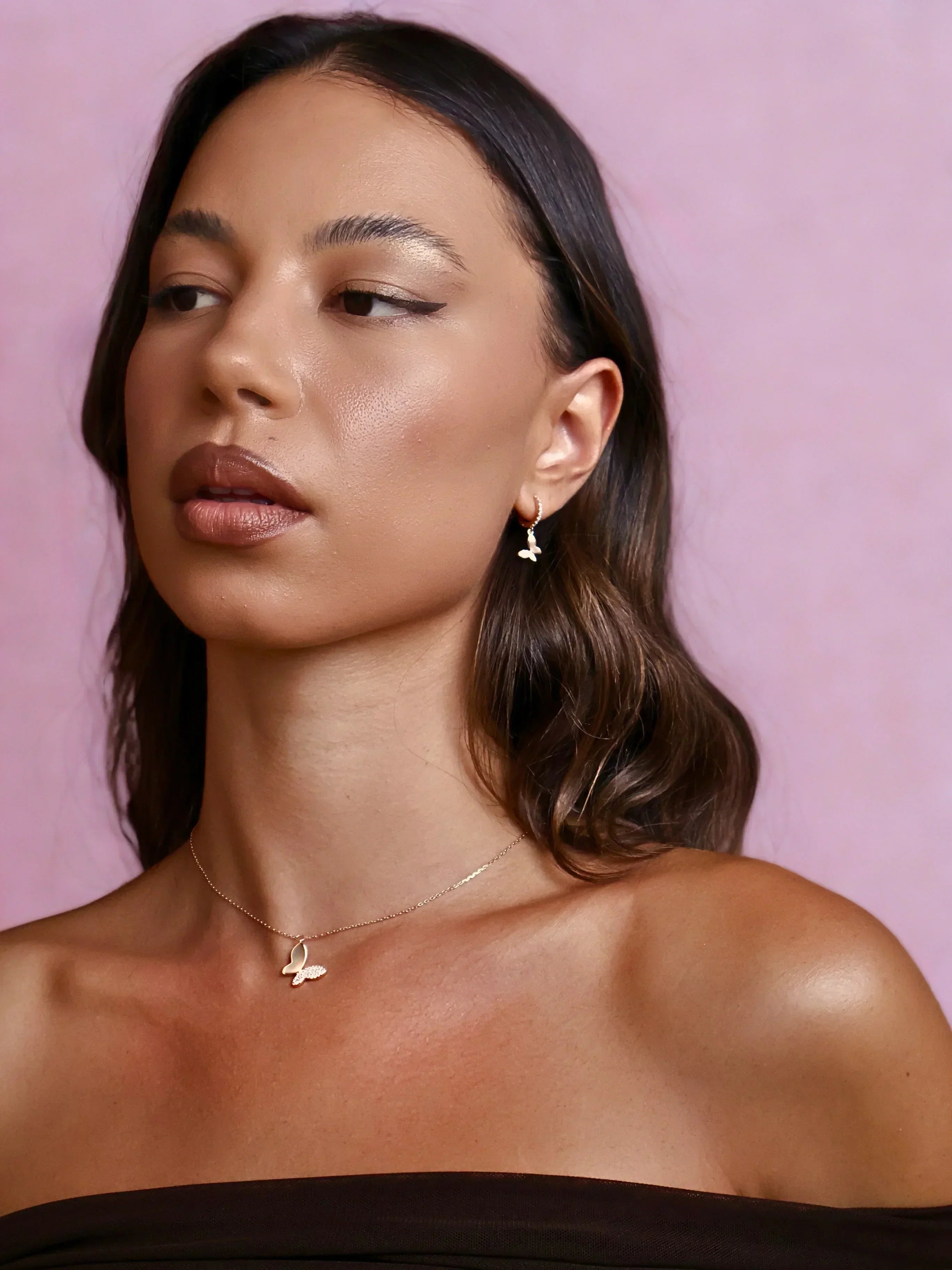 Close-up of woman with smooth skin wearing gold butterfly necklace and matching earrings against pink background