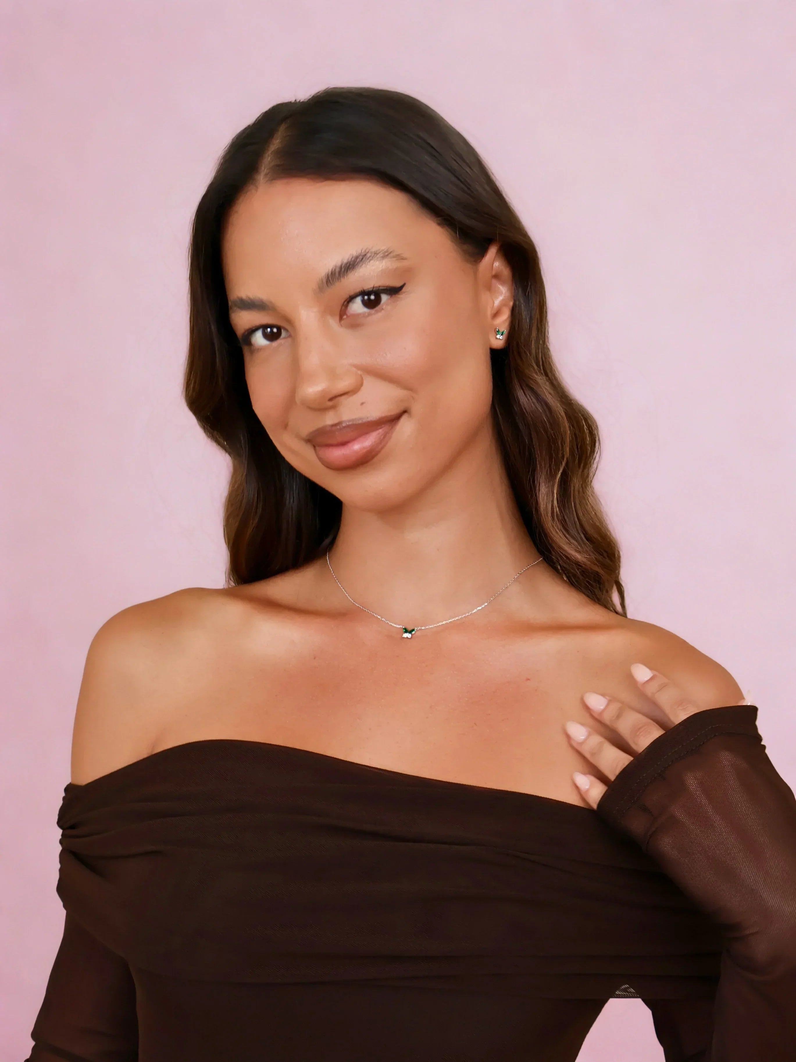 Woman wearing emerald gemstone necklace and matching earrings with off-shoulder brown top on pink background