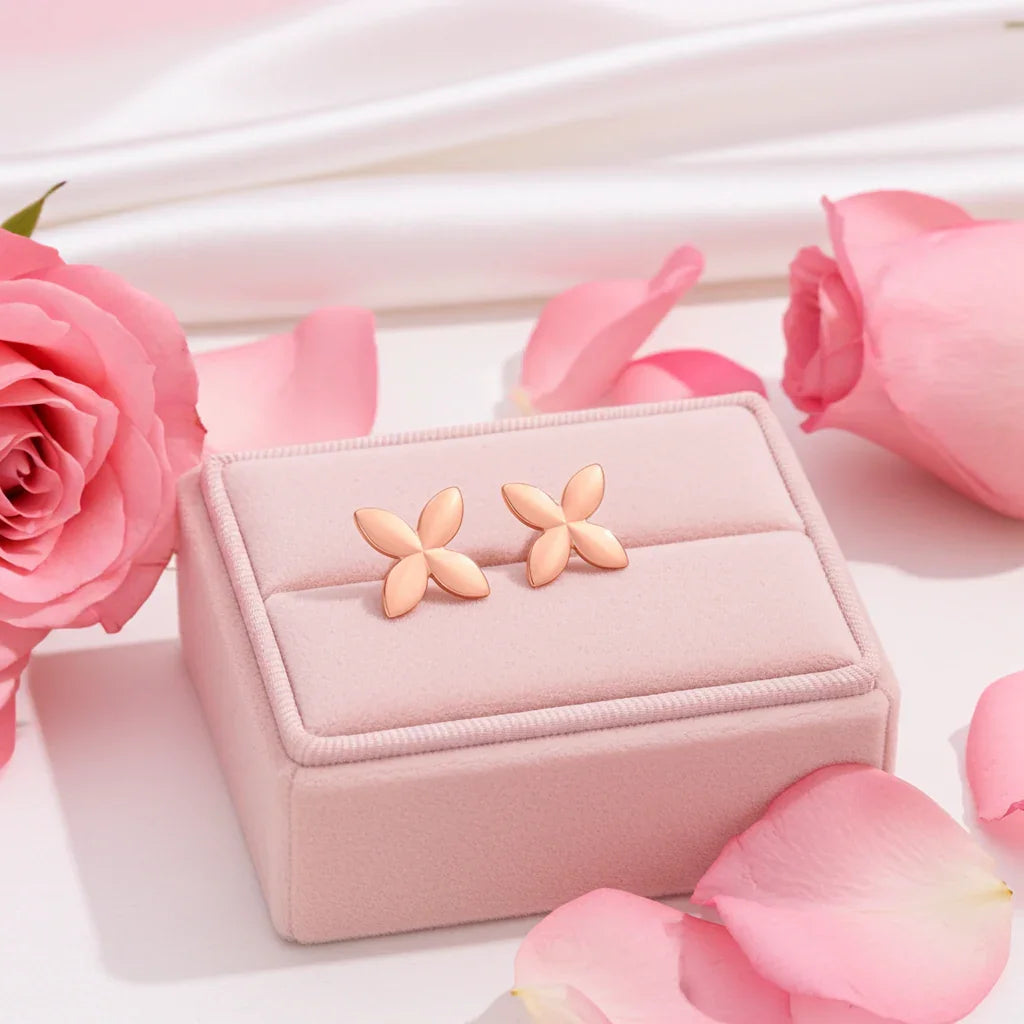 Rose gold minimalist butterfly stud earrings displayed on a soft pink cushion surrounded by pink rose petals and roses