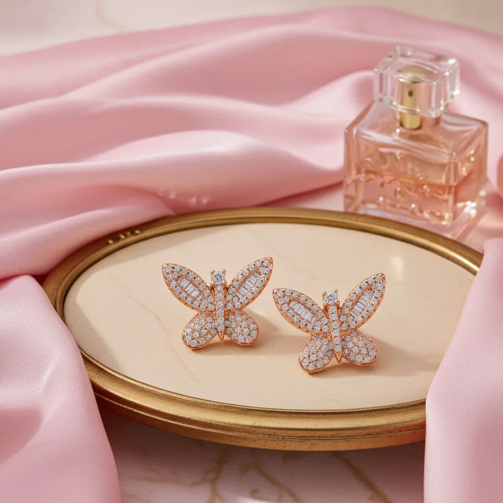 Rose gold butterfly-shaped earrings with sparkling crystals on a gold-trimmed marble tray with pink satin and perfume bottle