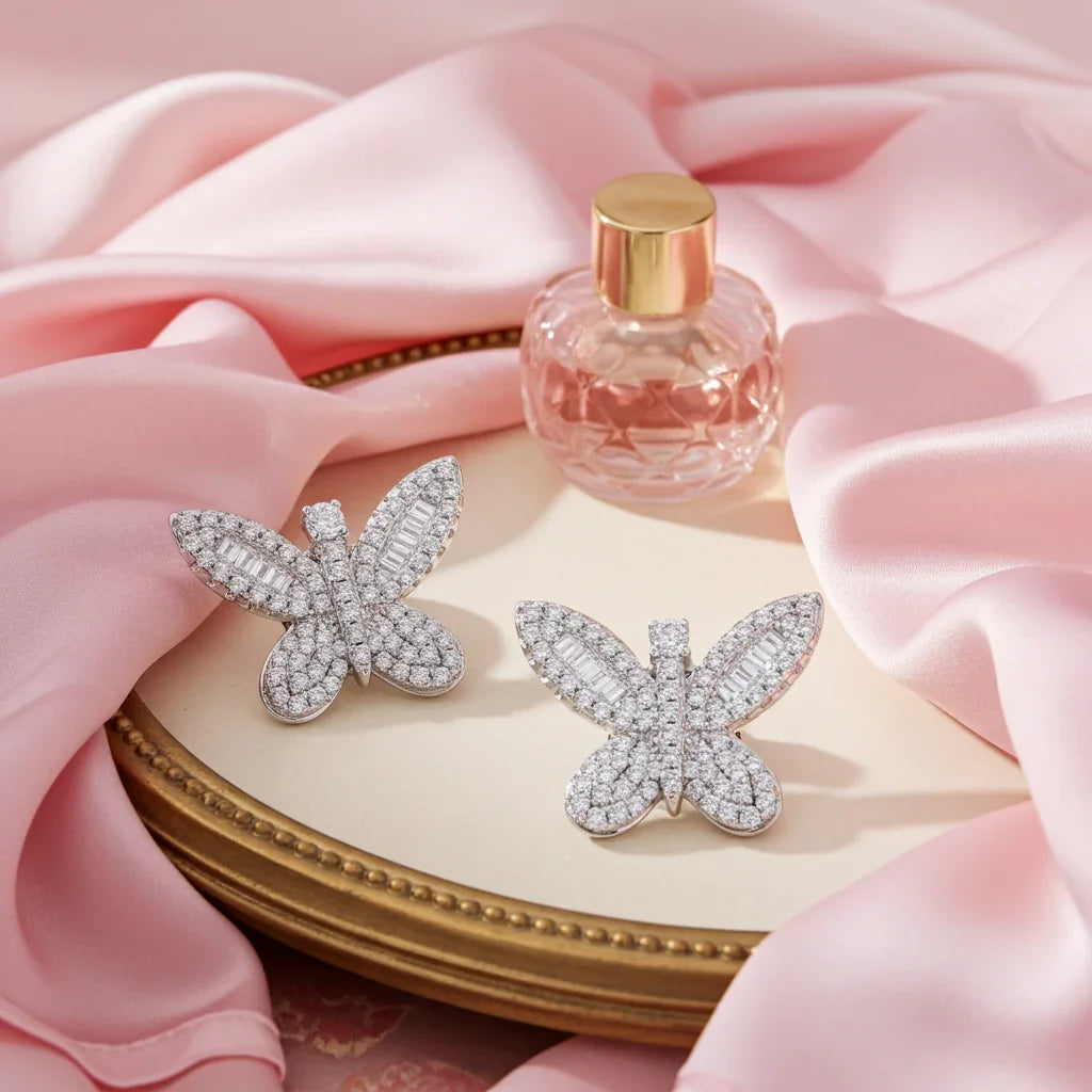 Silver butterfly earrings with sparkling crystals on gold tray, pink satin fabric and glass perfume bottle