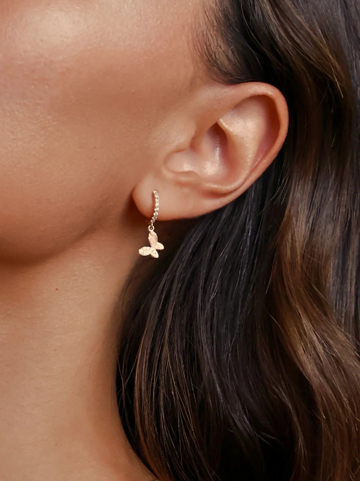 Close-up of woman wearing gold butterfly charm hoop earrings from Marwa Jewels with dark hair