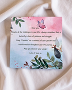 A square printed card with a watercolor butterfly and botanical illustration, placed on soft white fabric. The card contains a memorial-style message starting with 'Dear you,' and the background is a clean white textile surface.