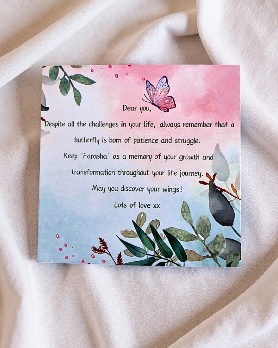 A square printed card with a watercolor butterfly and botanical illustration, placed on soft white fabric. The card contains a memorial-style message starting with 'Dear you,' and the background is a clean white textile surface.