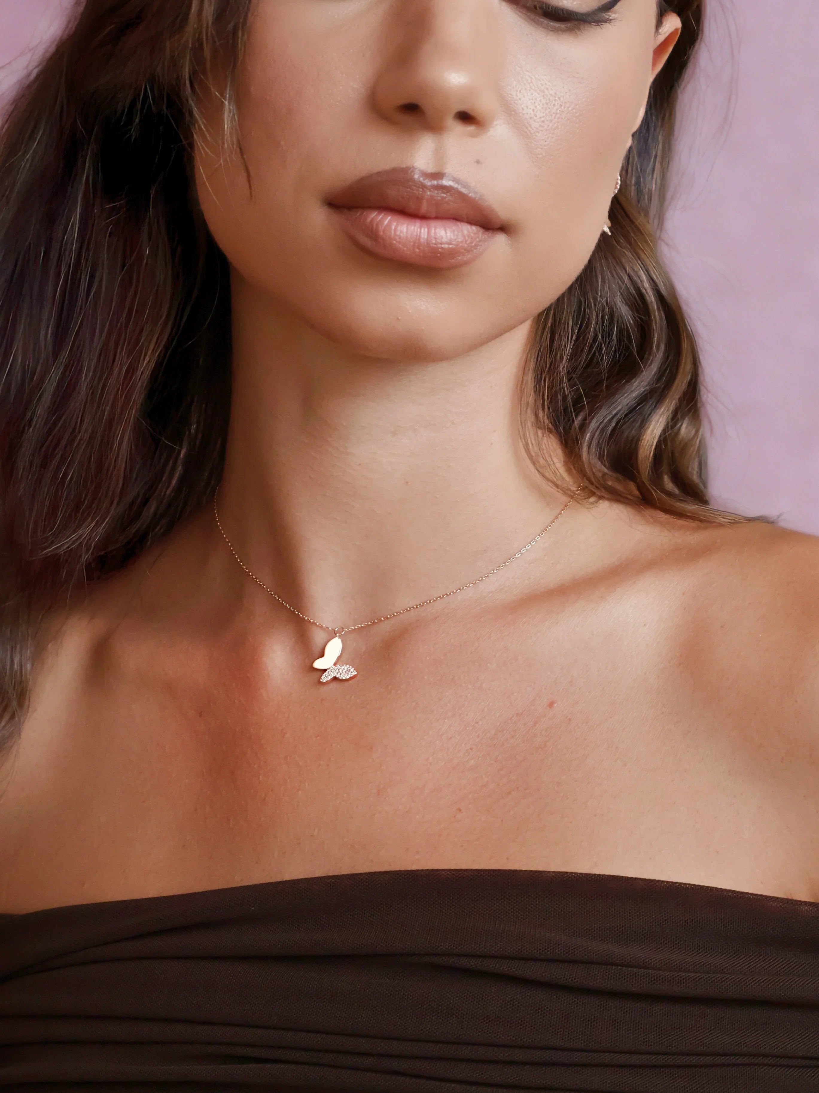 Close-up of woman wearing delicate gold butterfly pendant necklace and dark off-shoulder top against pink background
