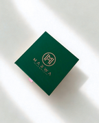 A dark green square product box with gold foil branding for 'MARWA JEWELLERY' and a circular 'MJ' monogram, photographed on a white surface with soft diagonal shadows.