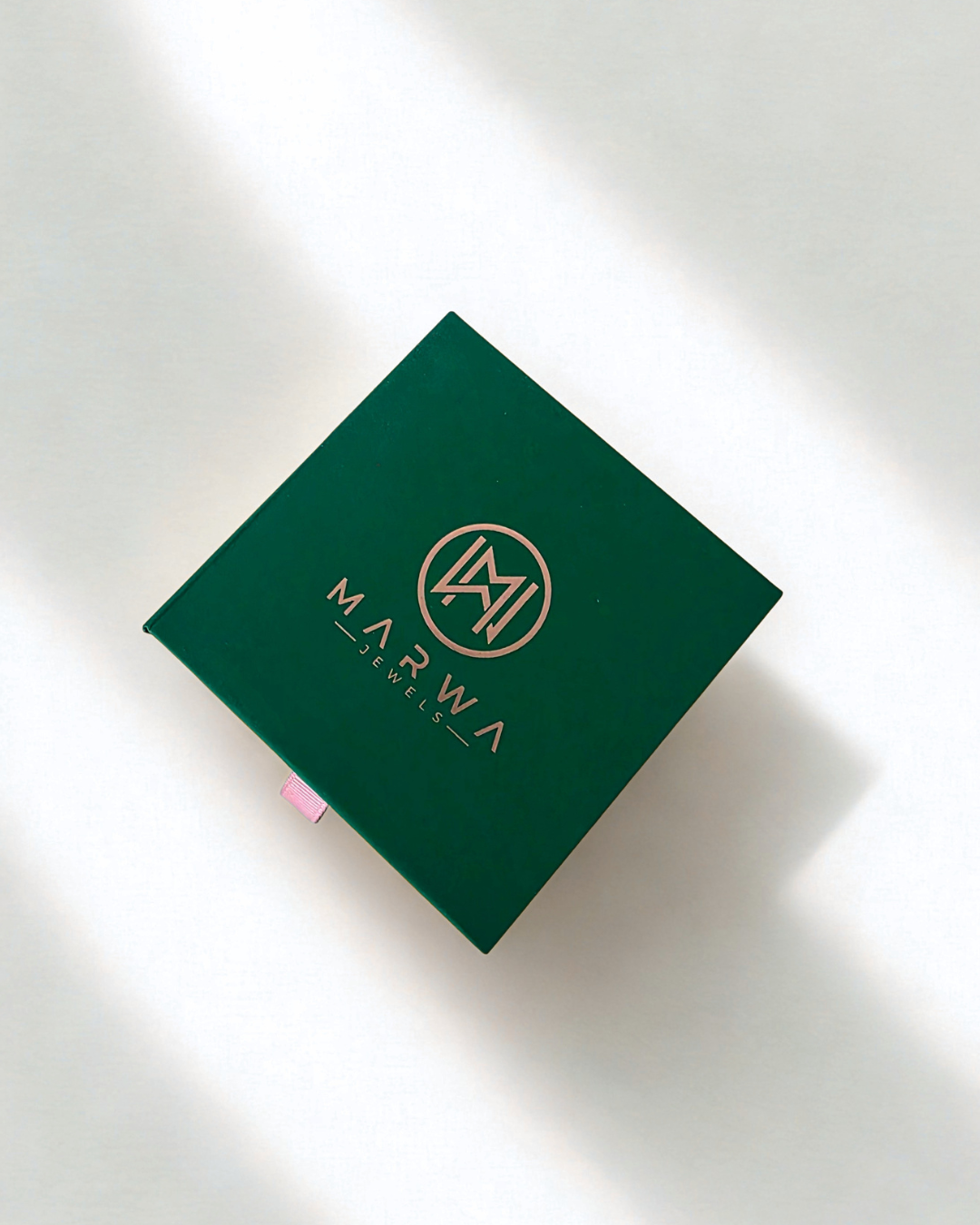 A dark green square product box with gold foil branding for 'MARWA JEWELLERY' and a circular 'MJ' monogram, photographed on a white surface with soft diagonal shadows.