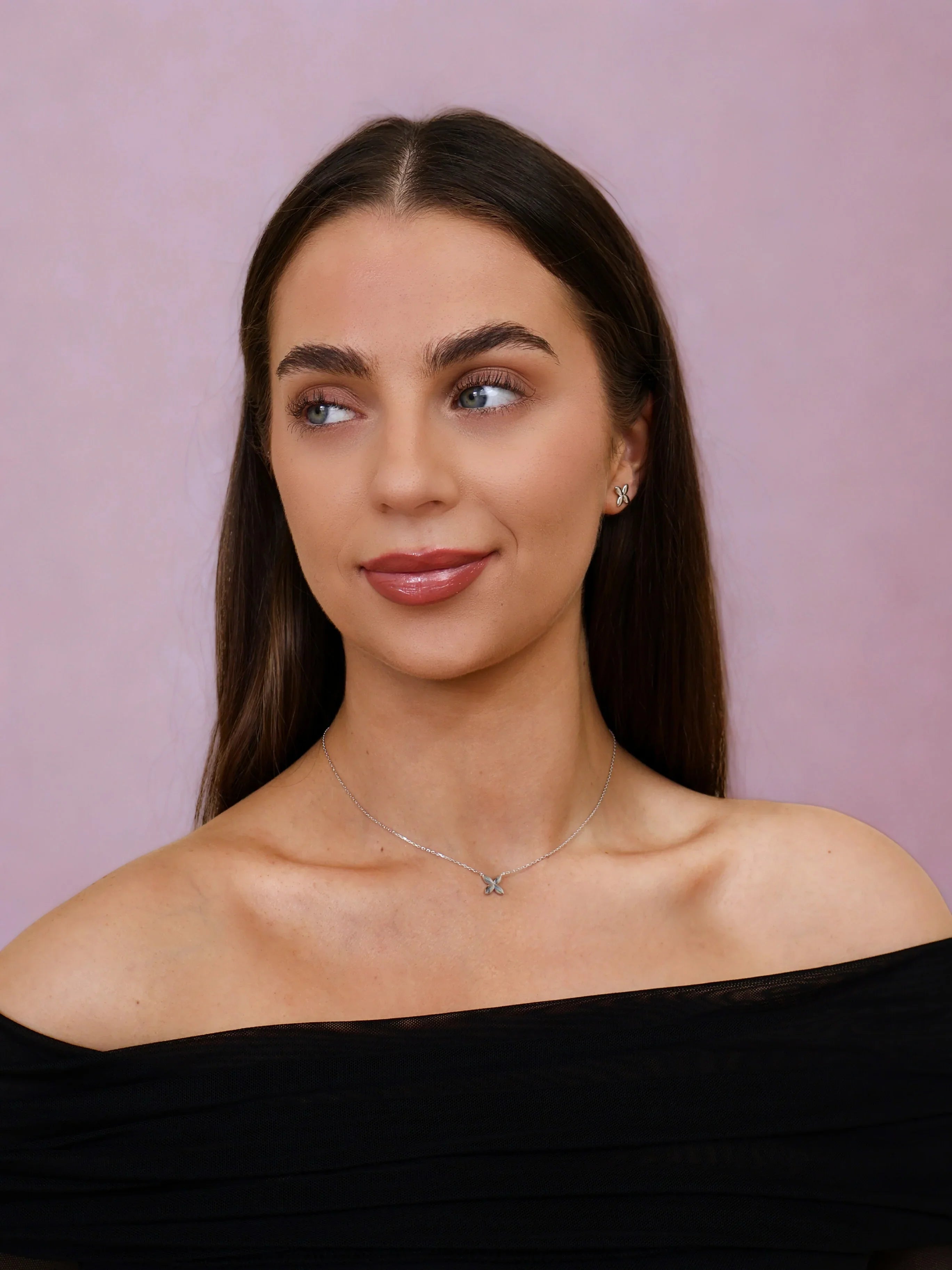 Woman wearing silver butterfly necklace and matching earrings with off-shoulder black top against pink background