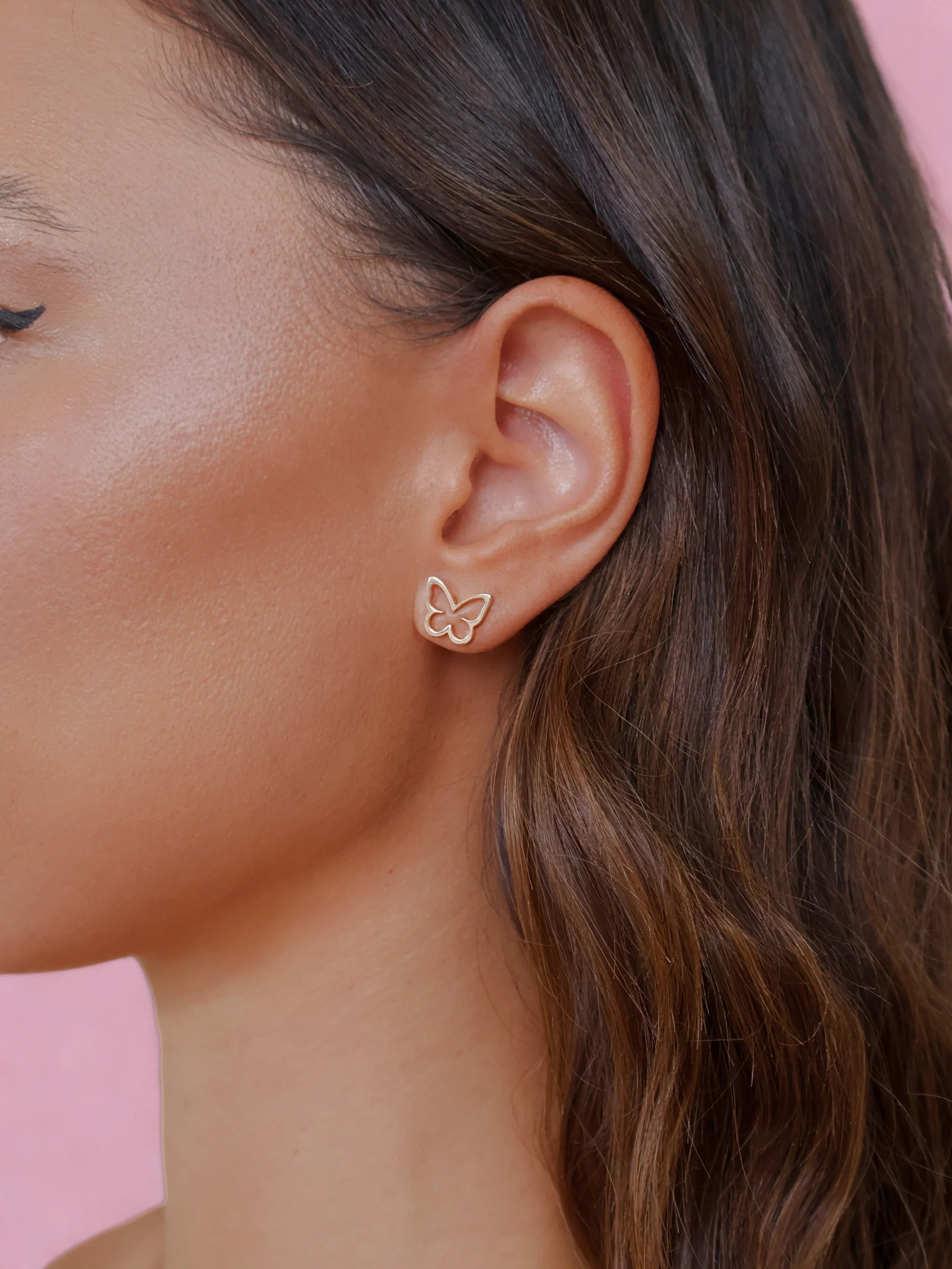 Close-up of a woman's ear wearing delicate gold butterfly stud earrings against a soft pink background